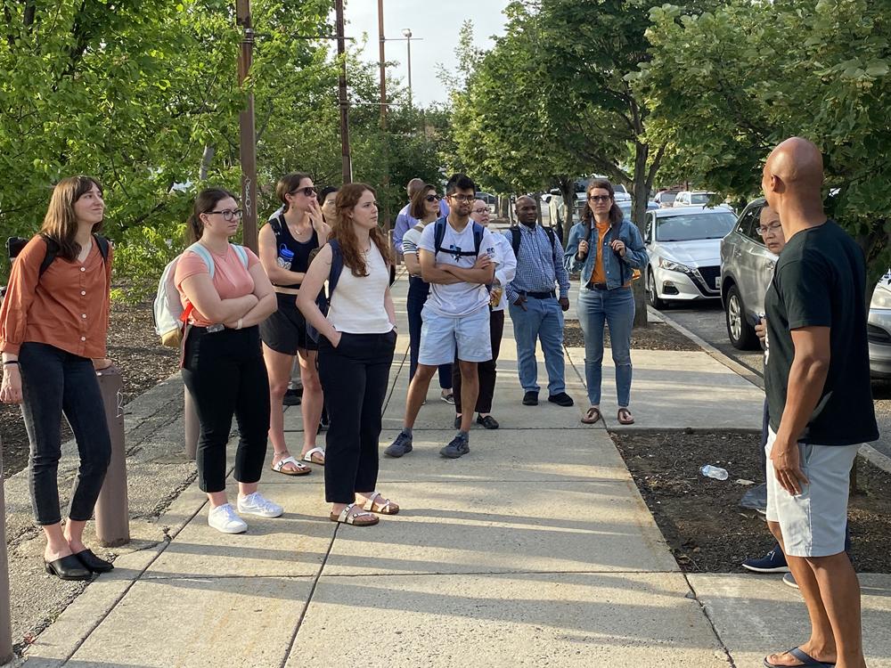 Ten researchers wearing backpacks stand on a sidewalk facing a man in a black shirt