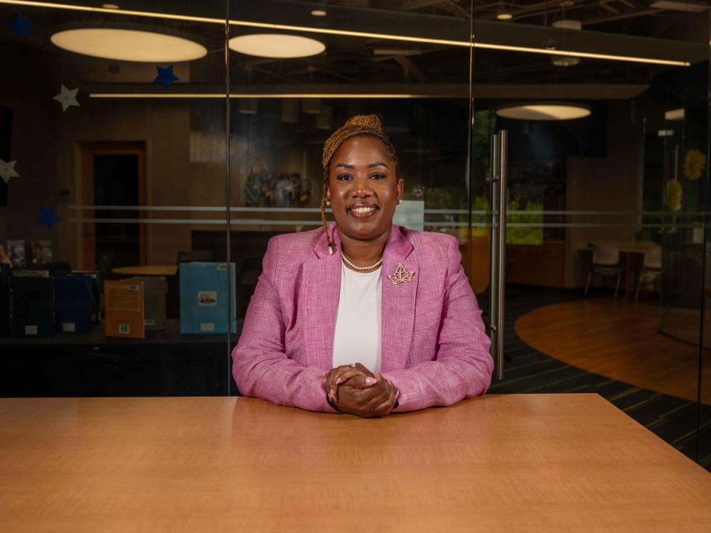 A smiling woman in a blazer sits at a desk with her hands clasped.