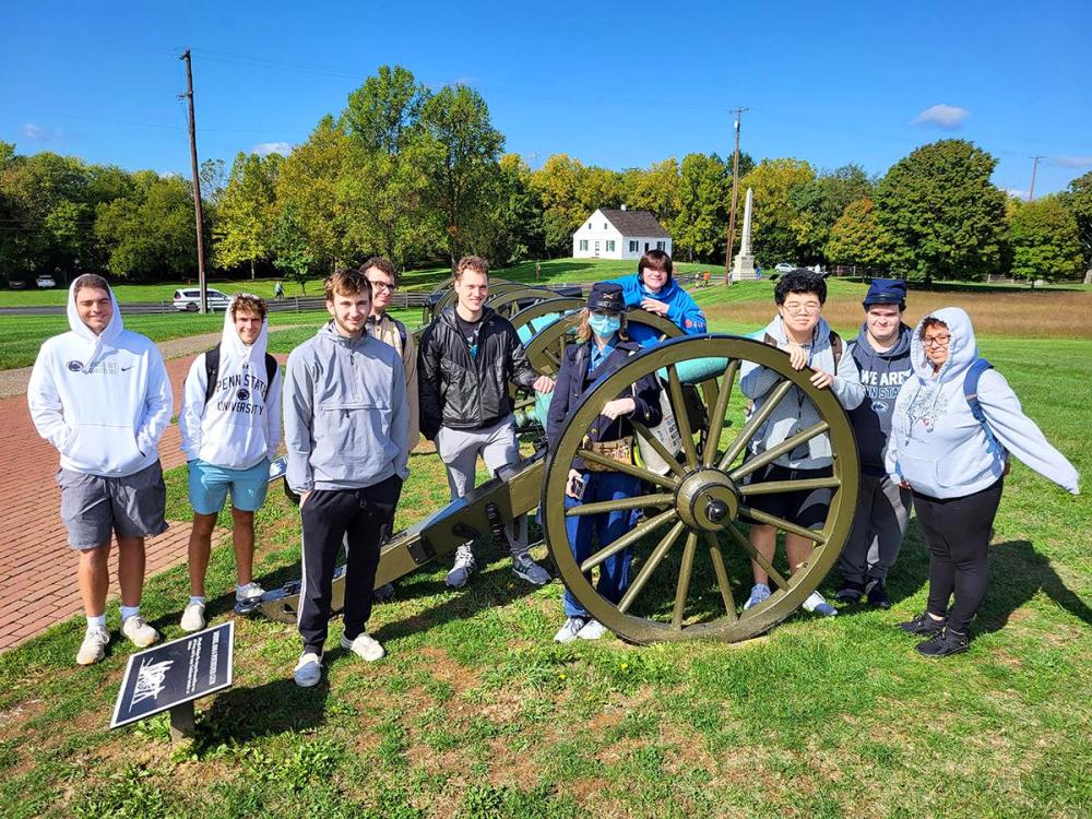 A group of students pose around a historic cannon on a sunny campus lawn.