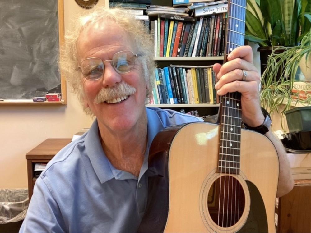 Photograph of a smiling man with glasses and mustache holding an acoustic guitar in a home office.