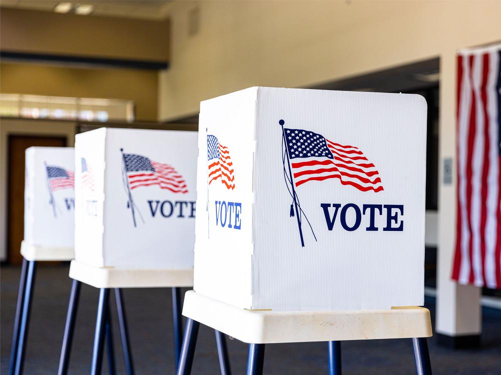 three empty election booths sit beside one another in a community center, each adorned with a graphic showing an american flag