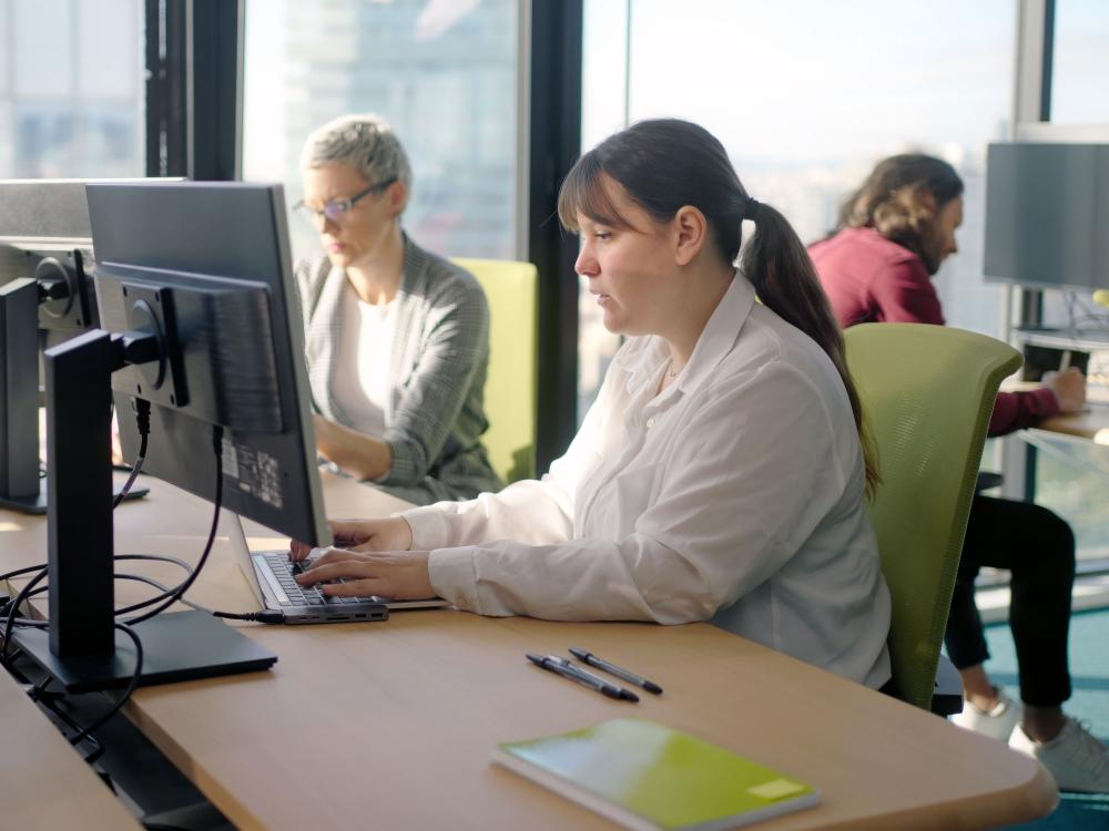 Three employees work at computers in a bright, modern office with large windows. In the foreground, a woman in a white blouse types on a laptop, while two colleagues work at separate desks behind her.