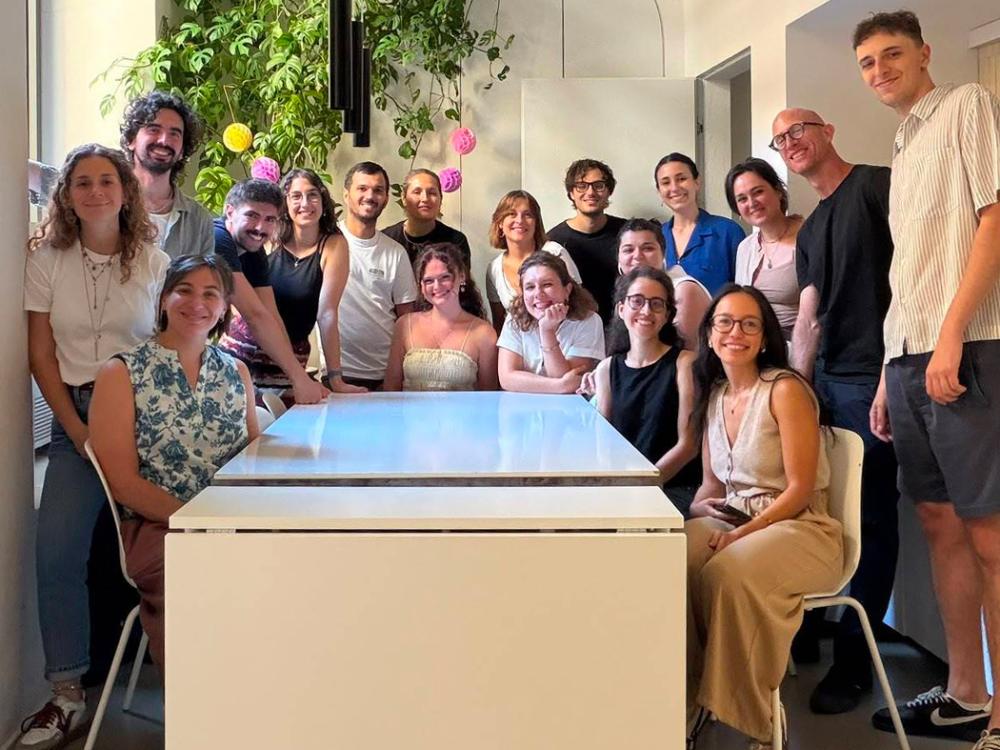 Group photo of 17 people smiling and posing around a large table in a bright room decorated with plants and colorful hanging ornaments.