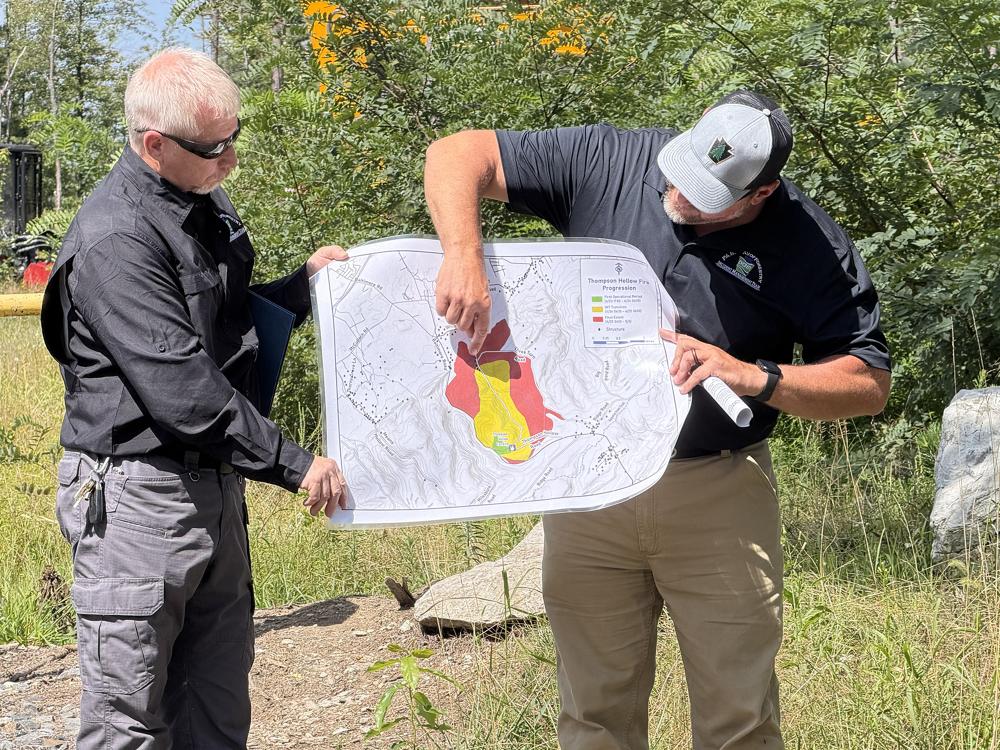 Two men in black shirts hold a paper map with red and yellow shaded areas in the middle. The man on the right points to the shaded region