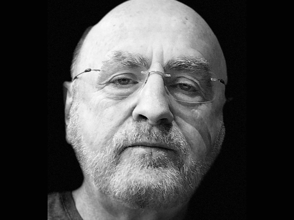 Black-and-white close-up portrait of an older bald man with a full white beard and wire-frame glasses, looking directly at the camera against a dark background.