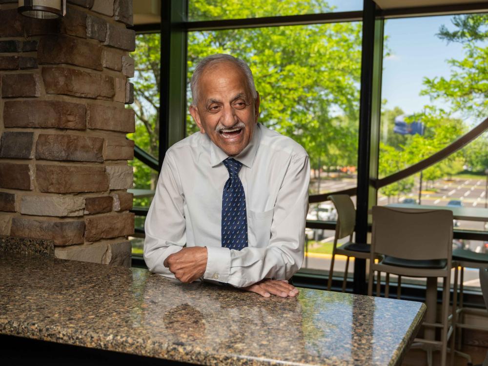 A laughing man in a button-down shirt and tie leans against a marble table in a lounge with trees outside.