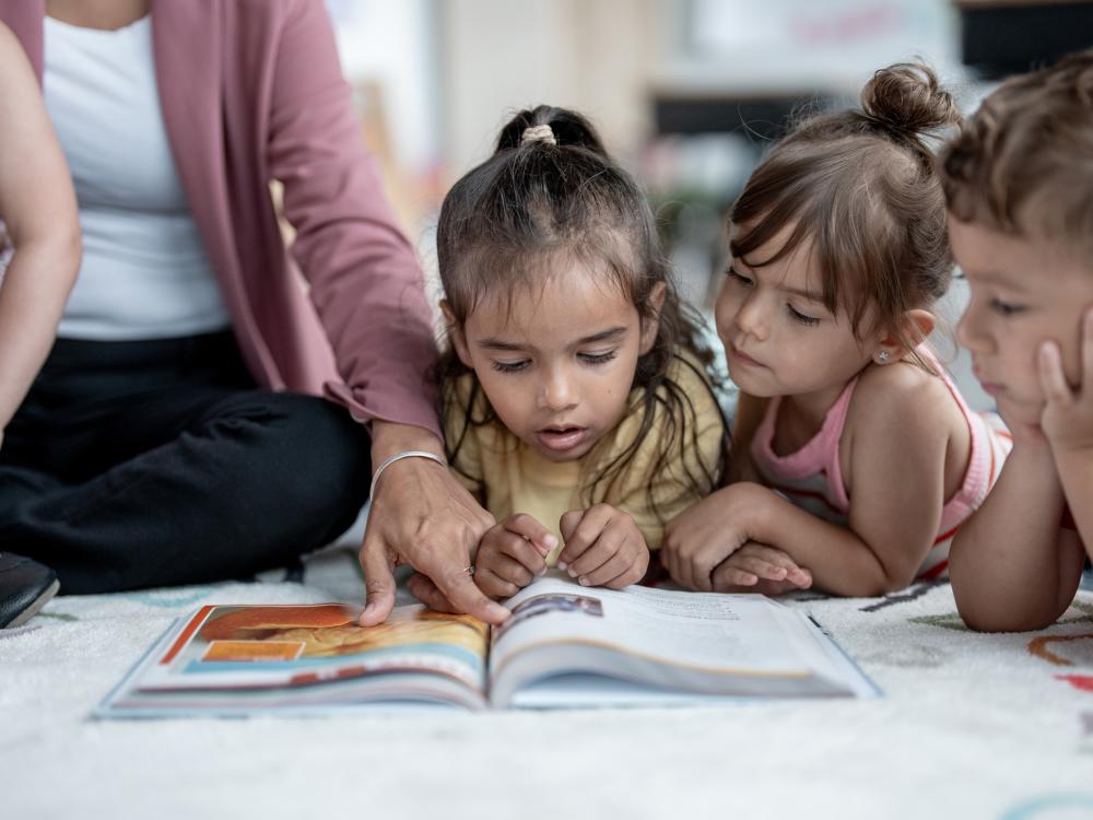 Preschool students gather around a book that their teacher is reading on a classroom floor.