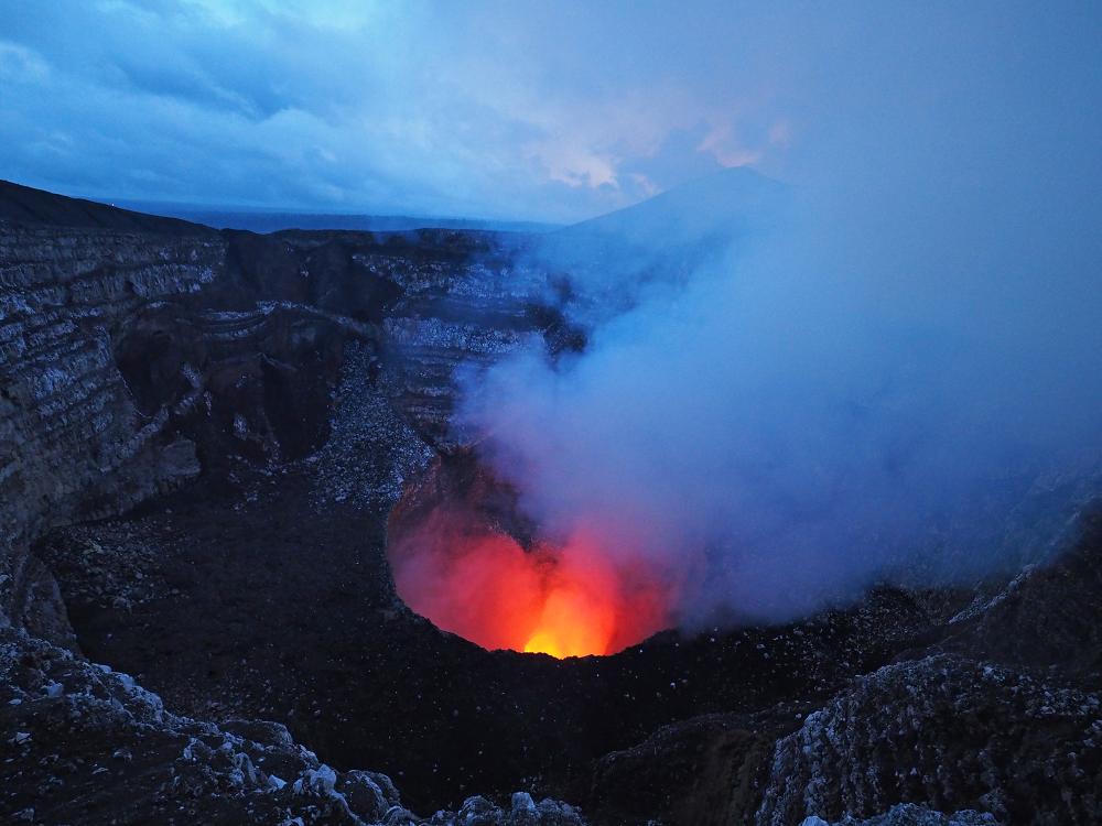 A crater lit with red and yellow sitting in a dark rocky landscape. Steam, appearing blue, emanates from the crater to the right.