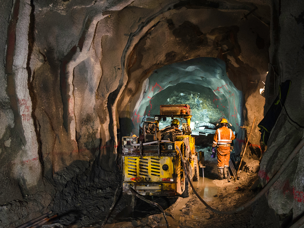 Mining workers and yellow drilling equipment inside an underground tunnel.