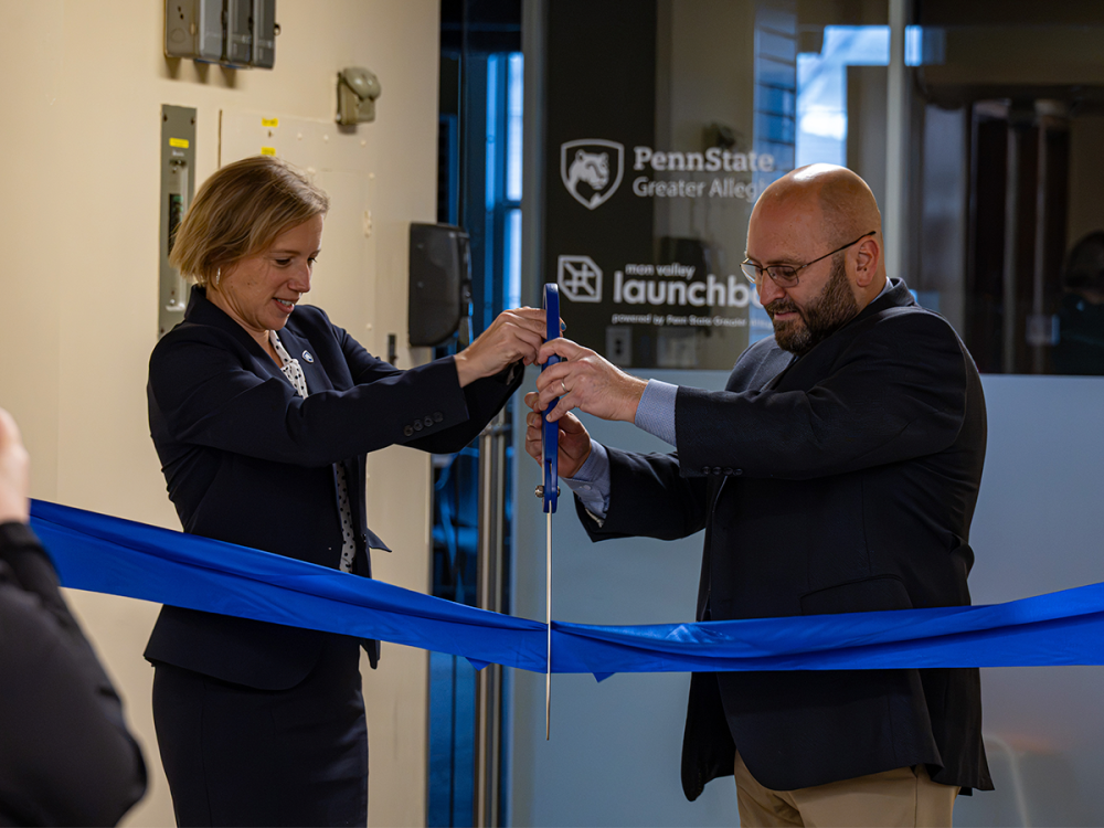 Megan Nagel, left, and Mayor Tom Maglicco hold oversized scissors over a blue ribbon, with Penn State Greater Allegheny and Mon Valley LaunchBox signage behind them.