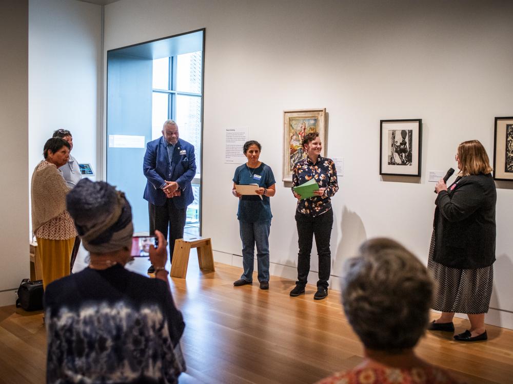 A group of people standing in an art museum. The person at right holds a mic and is introducing the three people to her left.