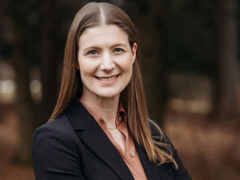 Rachel Shelden wears a dark blazer and orange blouse and stands against outdoor backdrop