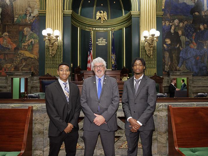 Three people stand in a formal legislative chamber with golden columns, historic murals, and United States and Pennsylvania flags behind them.