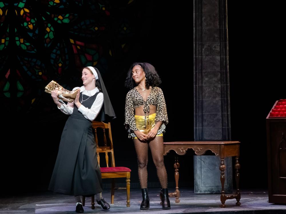 A performer dressed as a nun and a performer dressed in street clothes perform on stage
