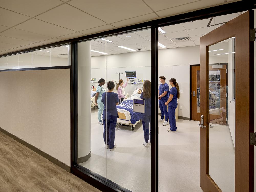 Nursing students gathered around a hospital bed in a simulation classroom.