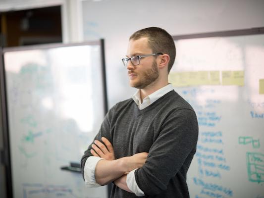 A professor stands with his arms crossed in front of whiteboards. 