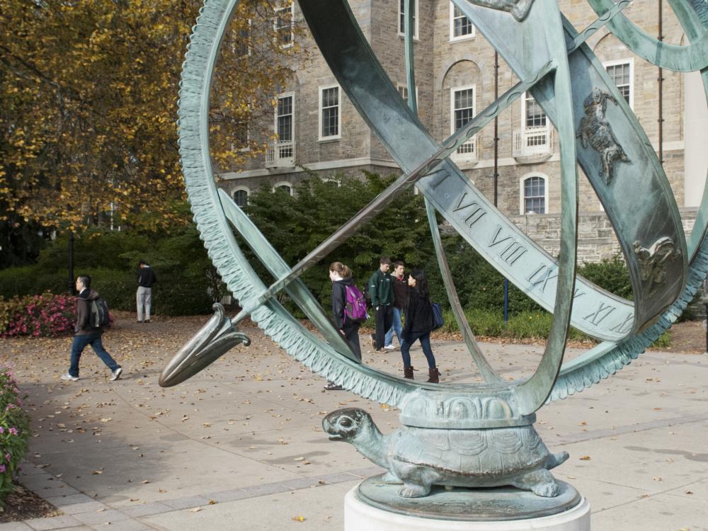 The armilllary sphere rests on the back of a turtle, with students walking by on the sidewalk on a fall day