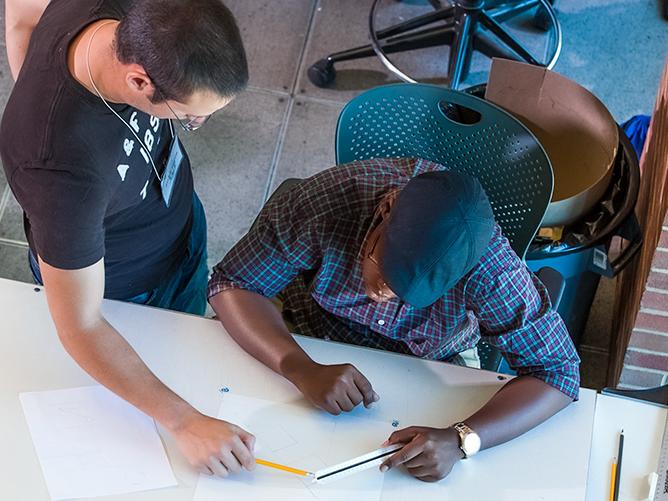 A faculty member instructs an architecture student as he sits at his drawing table.