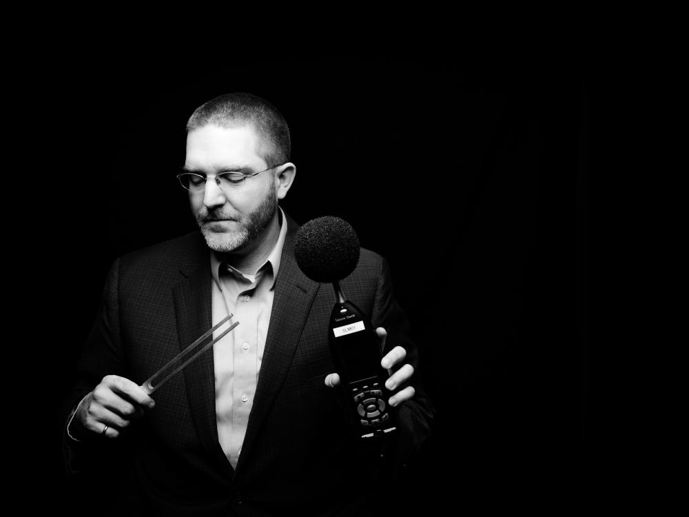 black and white photo of a professor posing with acoustics equipment (tuning fork and microphone)