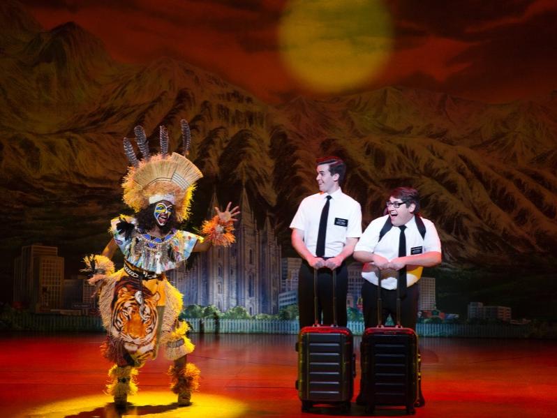 A man dressed in garb made from straw, feathers and beads waves his hand as he sings while two man dressed in similar outfits of pants, a button-down shirt, and tie and holding on to luggage watch and smile.