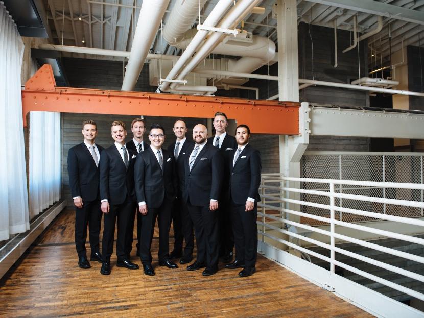 A group of men wearing the same suits stand in a cluster in a chic industrial surrounding.