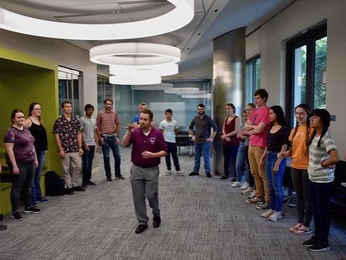 Man in red shirt standing in center of circle of students and gesturing with hands.