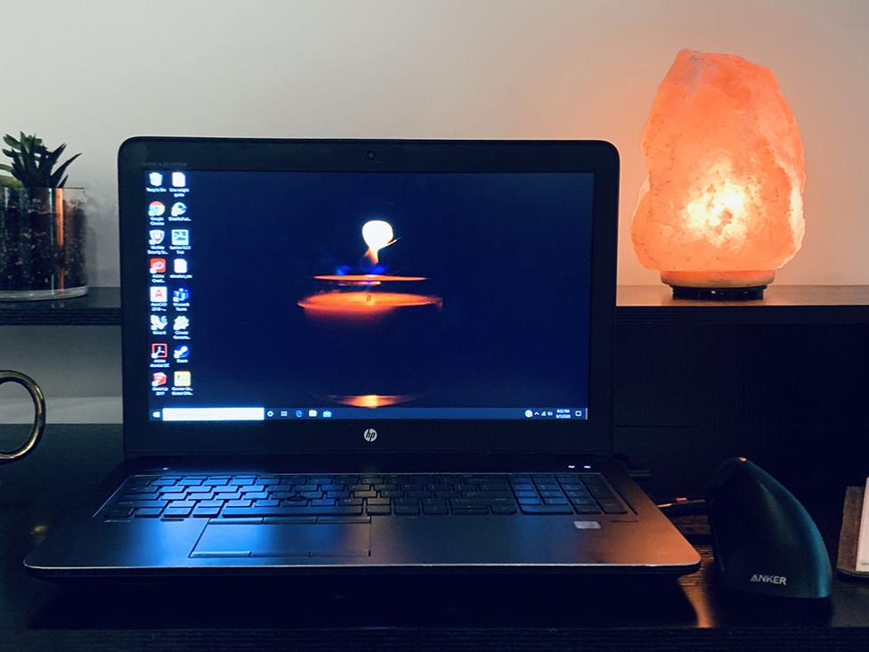 Photo of a home workspace with a laptop, coffee mug, Himalayan salt rock lamp, a water bottle, a sketch pad, and a plant.
