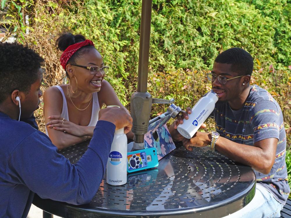 Three students sitting around a circular table outside. One student is holding their white water bottle displaying Greater Allegheny Logo 