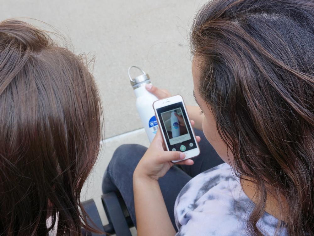 Two students sitting. One student is holding a phone while scanning a barcode that is on her water bottle