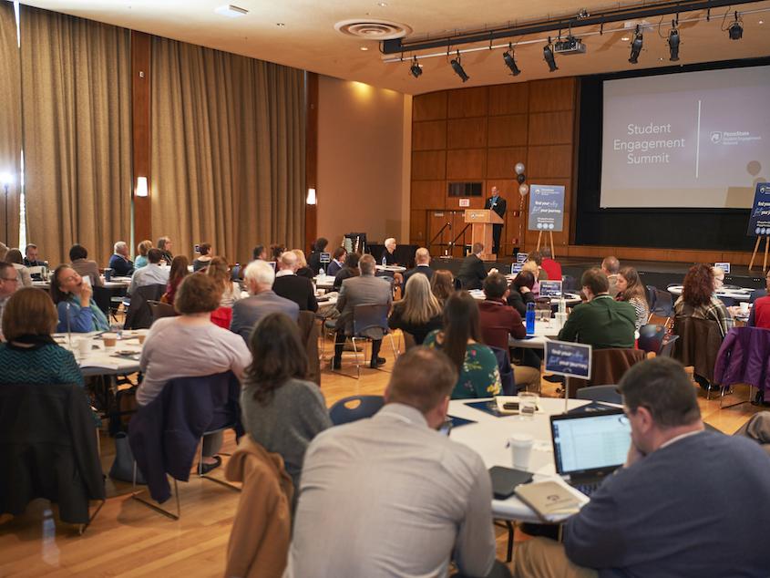 An audience sits at tables in the HUB Alumni room during presentation