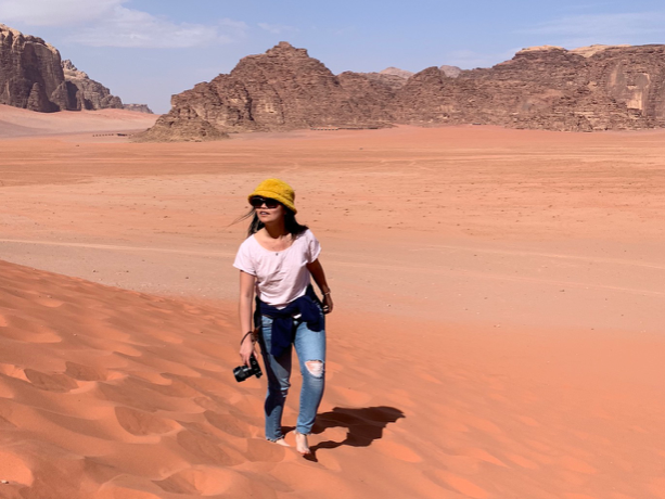 Young woman wearing sunglasses, a yellow hat, white t-shirt and jeans walks through the desert with a camera and stares off into the distance.