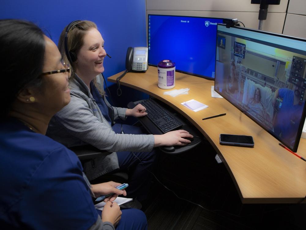 Jennifer Gish, nurse manager, and registered nurse Marilou Magnaye sit at a curved desk and smile at a computer monitor that shows patient Kimberly Laughman in her hospital room in bed, with nurses and equipment around her. 