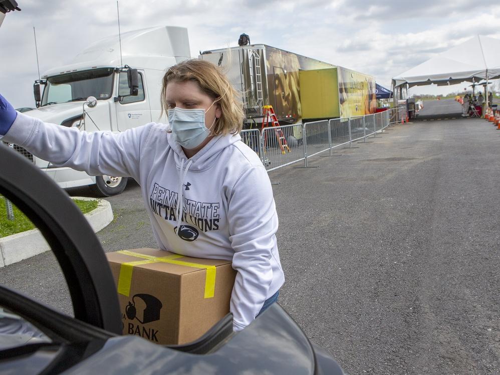 Registered nurse Stephanie McGaw, who is wearing a sweatshirt, gloves and a mask, lifts a cardboard box of food into the trunk of a car. A tractor-trailer truck and traffic cones marking drive-through lanes are in the parking lot behind her.