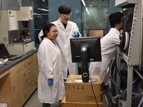 Two men and a woman in lab coats looking at polymer samples via a glove box and monitor.