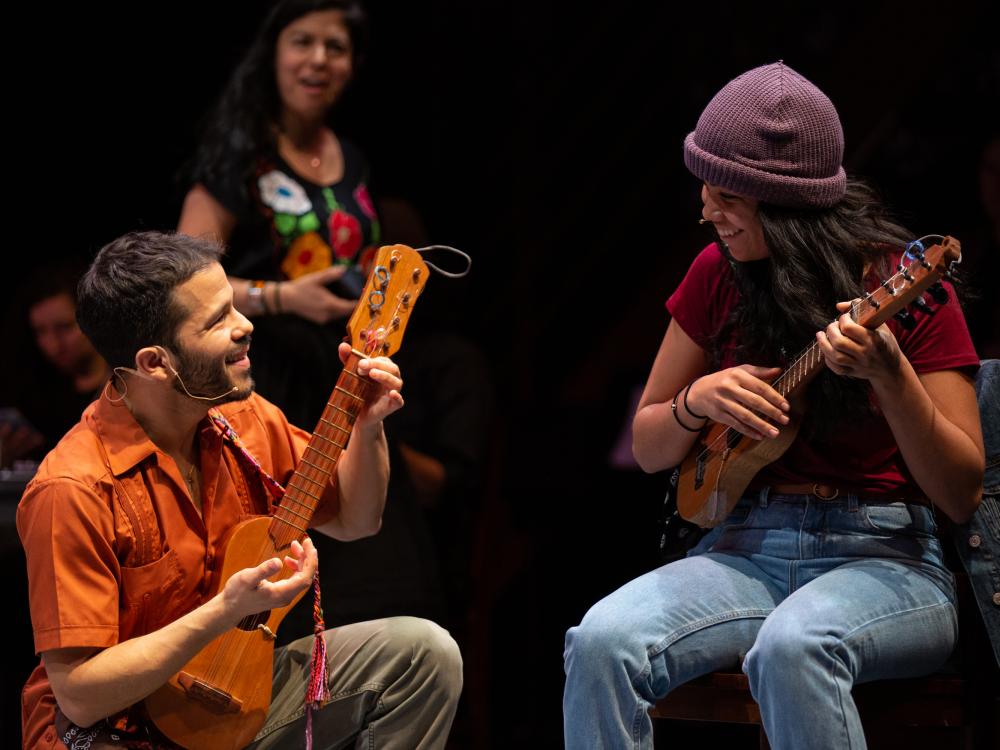 A man kneels next to a woman sitting in a chair while they both play stringed instruments.