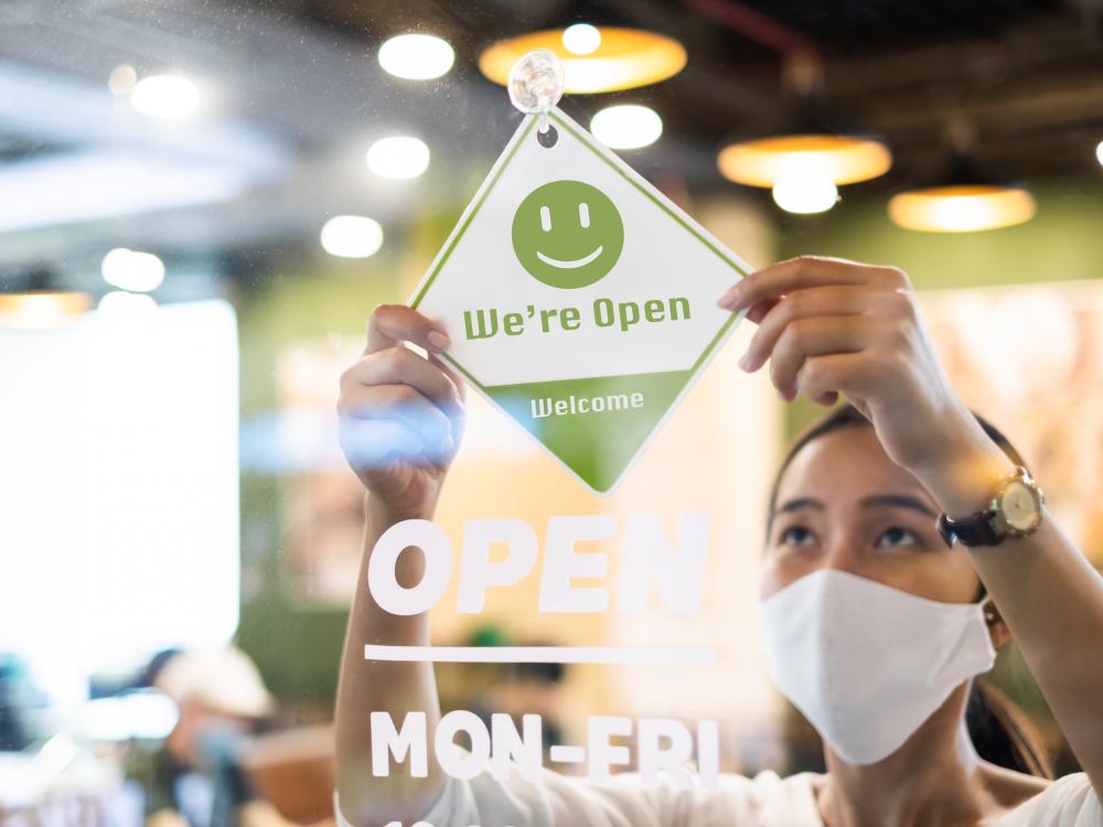 Woman in white mask hanging Open sign in restaurant window