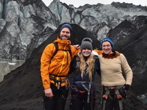 Three people standing on dark, rocky ground with rock formation behind them.