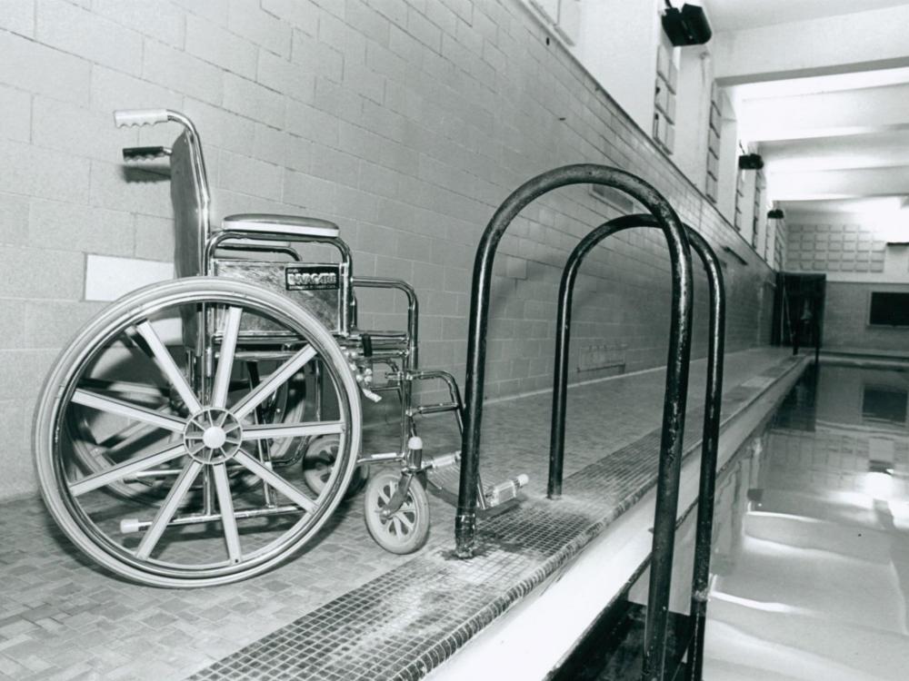 A black and white photo of a wheelchair next to a ladder leading into an indoor pool