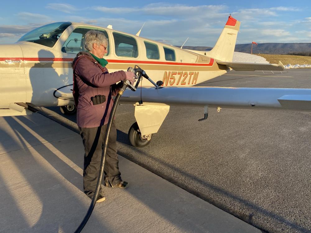A person holds a fuel pump to a small aircraft. A mountain is in the background. 