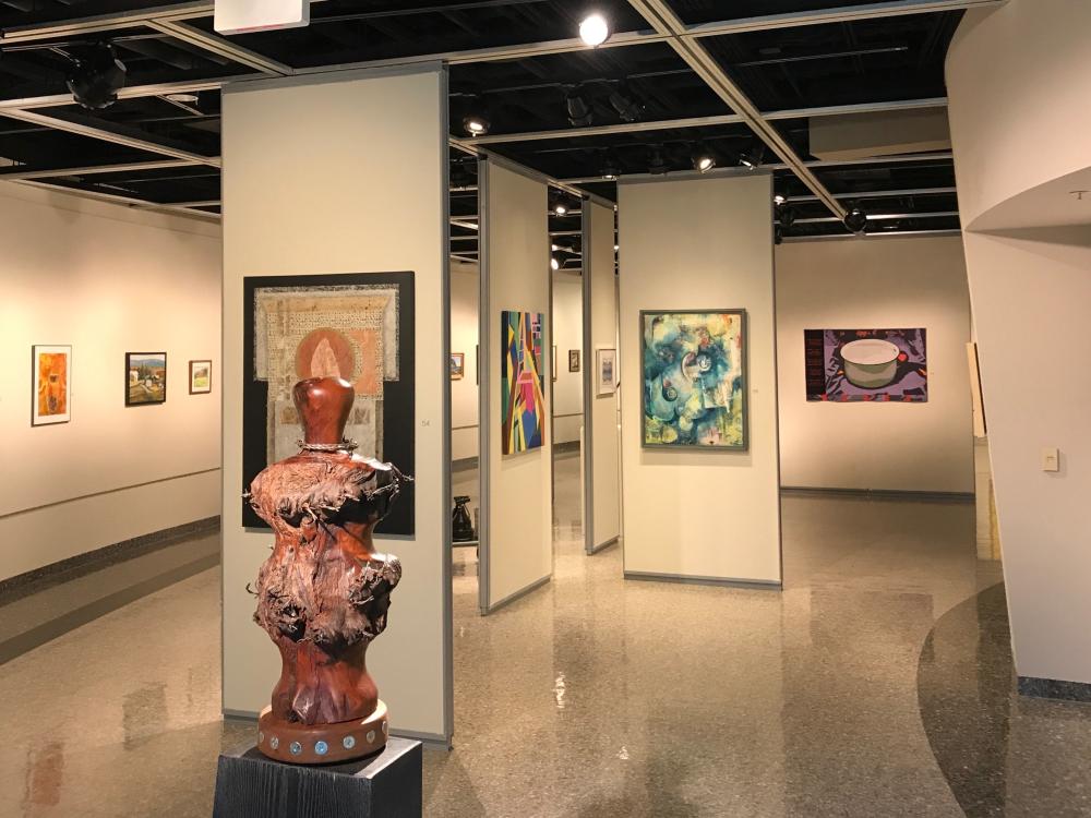 An overall photo of the Art Alliance exhibition in Robeson Gallery, showing a variety of paintings hanging on the walls with a large wooden sculpture in the foreground. 