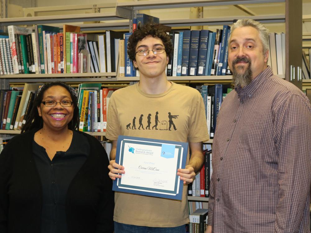 Three people standing in front of a bookshelf smiling. The person in the middle is holding an award. 