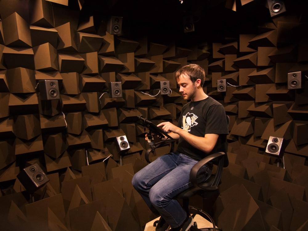 A student sits in a soundproof room surrounded by loudspeakers. 