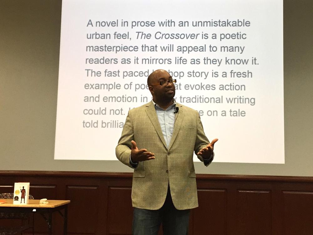 horizontal photo of Kwame Alexandar speaking to auditorium audience with review of his book "The Crossover" projected on wall behind him