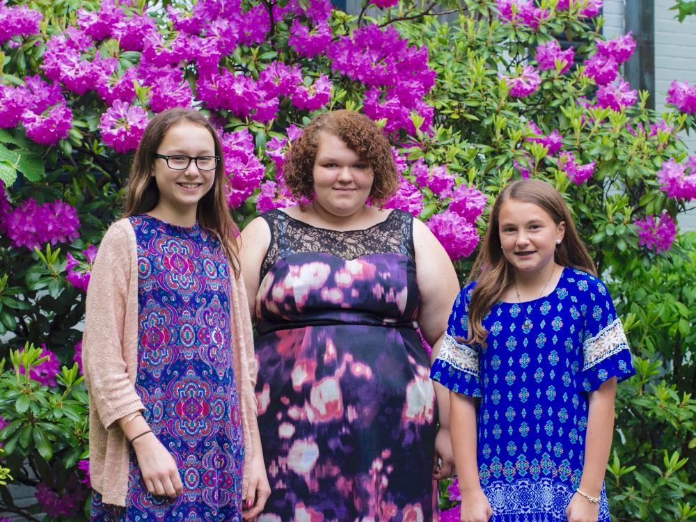 horizontal outdoor photo of three white female students standing in front of large blooming rhododendron shrub 