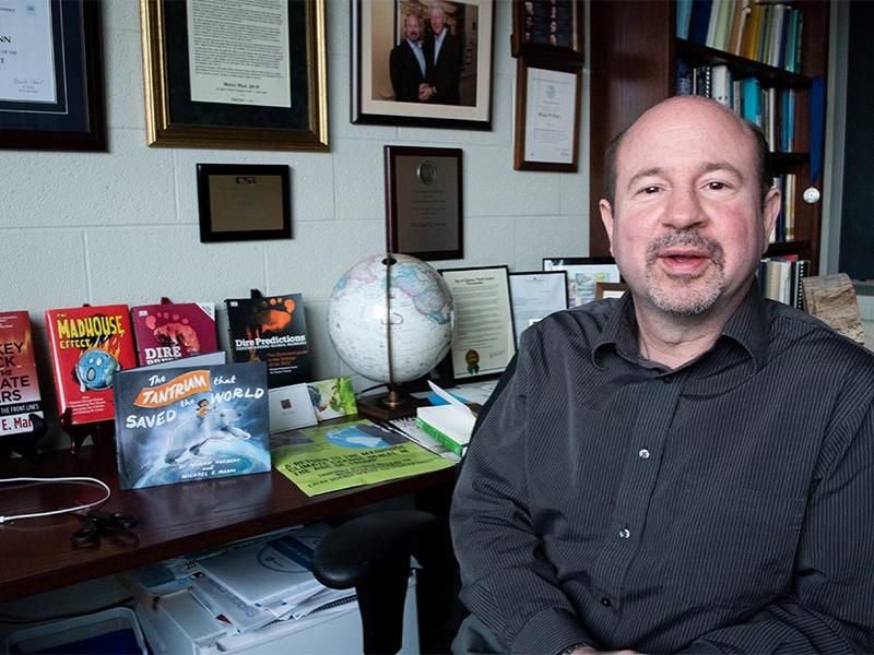 Michael Mann sitting in front of a table holding his many books on climate science.