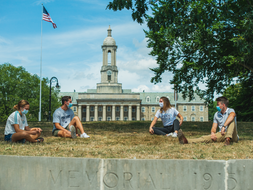 Four students sitting on the ground wearing masks with Old Main in the background on a clear day