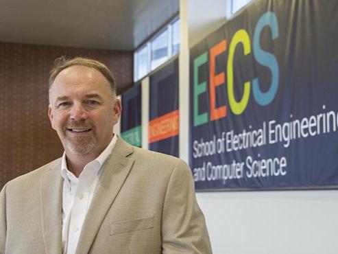 A man in a tan suit jacket poses in front of a School of EECS wall banner