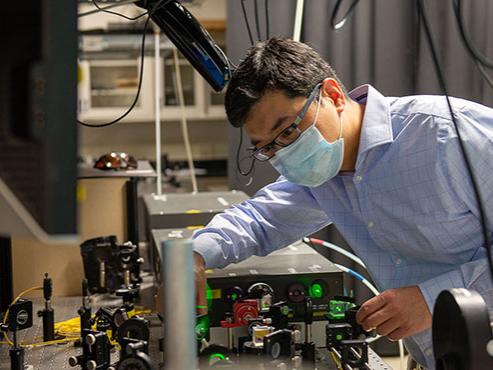 A man in a blue button-down shirt and a blue face mask adjusts optical devices that are shining green lights 