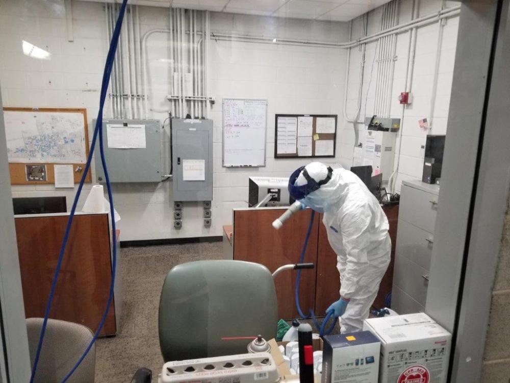 A member of OPP in personal protective equipment spraying disinfectant in a room on campus