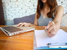 Woman seen from chin down is seated at wooden table. She has one hand on a laptop. Her other hand holds a pen which is poised above a clipboard with paper in it. 
